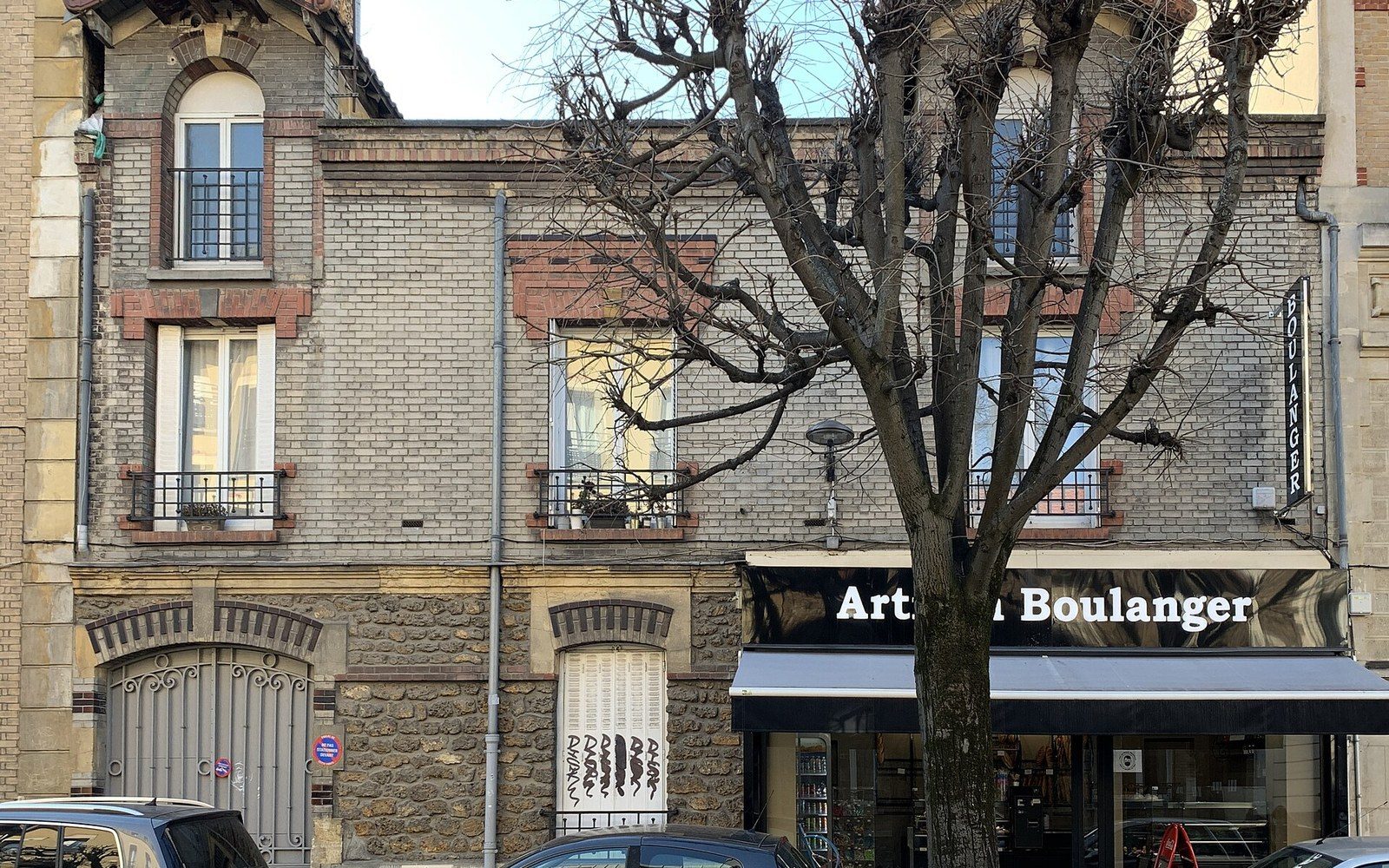 A bare tree stands before a brick building with a boulangerie storefront on the ground floor